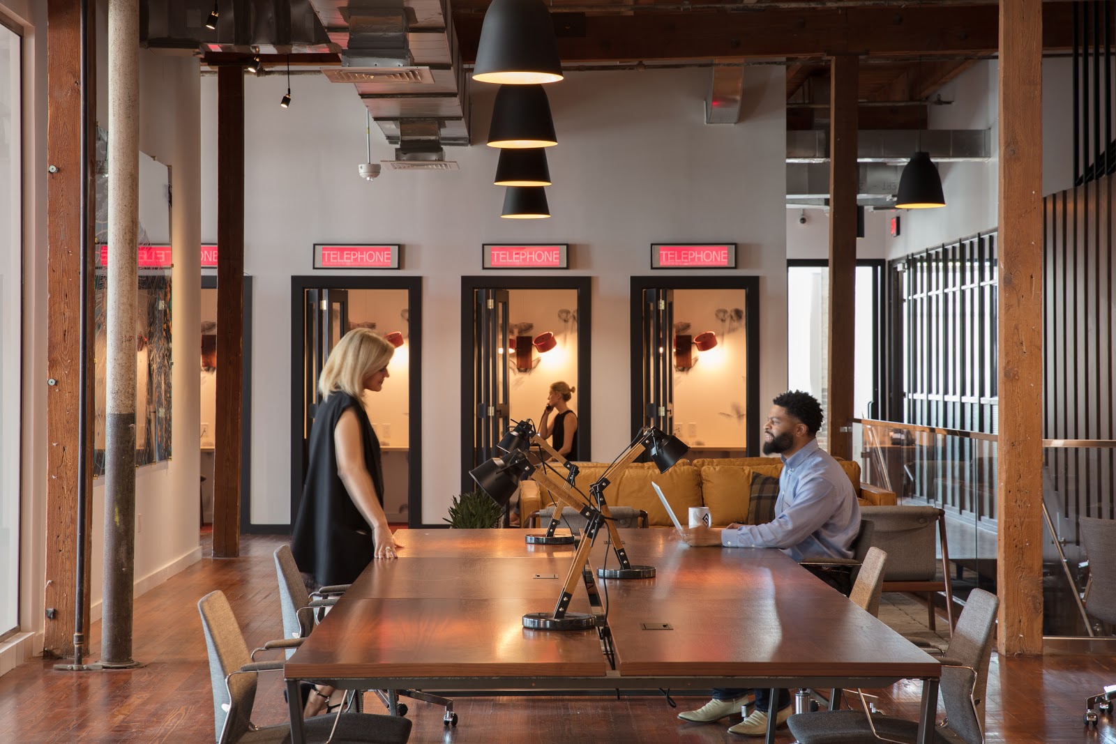 The Shop Coworking industrial workspace with dark wood communal table, black pendant lights, and three Telephone-labeled phone booths with red neon signs