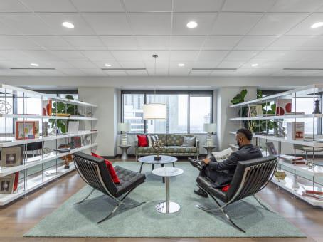 HQ Place St. Charles executive lounge with black Barcelona chairs, green area rug, open metal shelving, and member reading by city-view windows