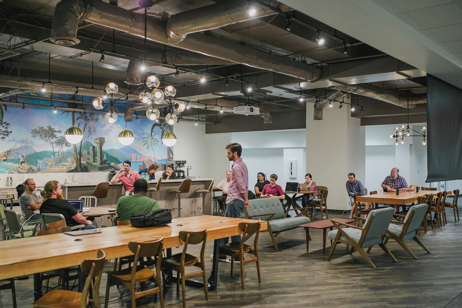 Launch Pad open event space with tropical mural wall, chrome bubble chandeliers, communal wood tables, and group meeting in progress