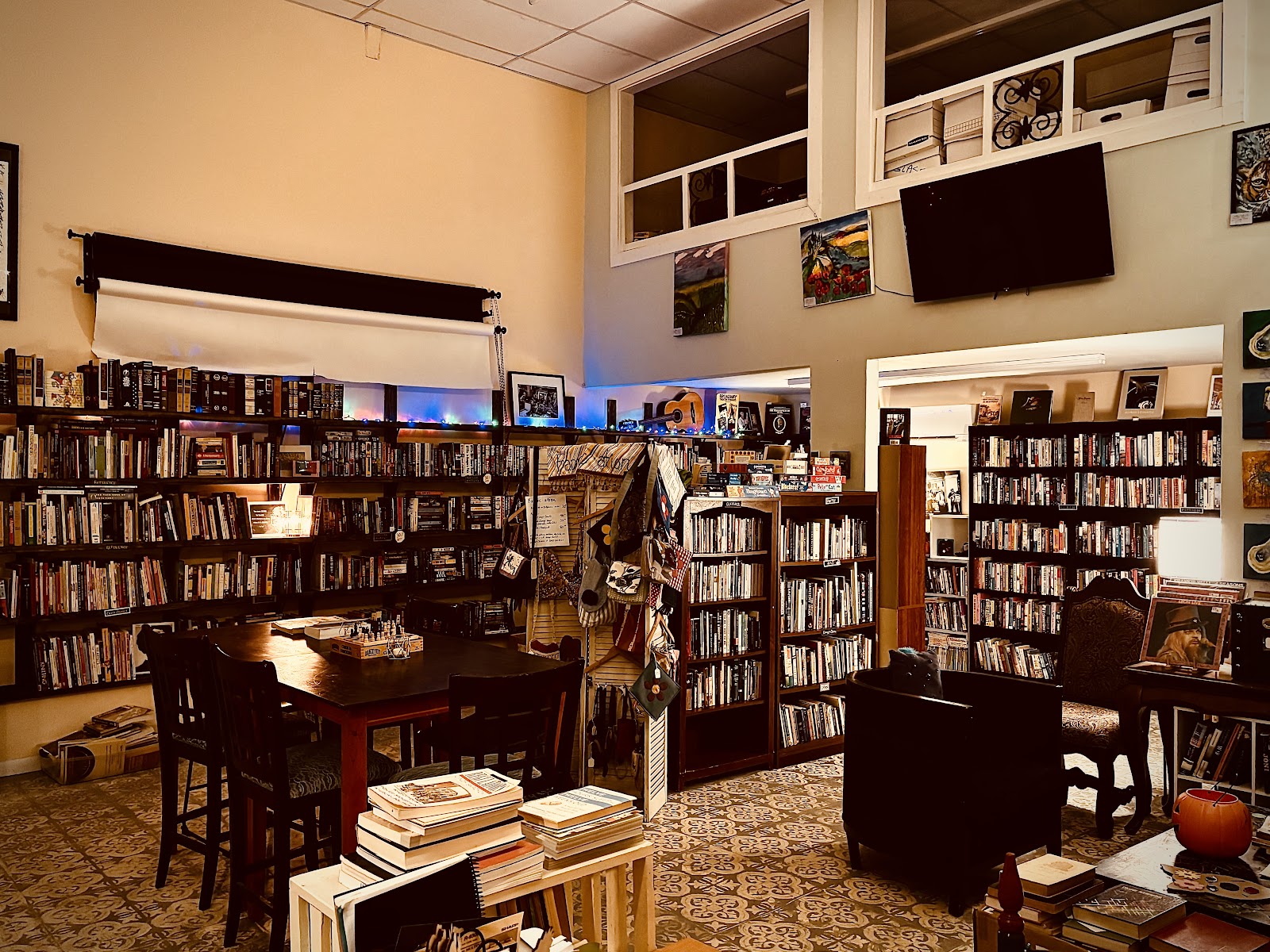 Nomad Used Bookstore interior packed with floor-to-ceiling bookshelves, dark wood table with chess board, accent lighting, and eclectic art